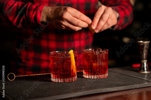 Bartender preparing two red Negroni style cocktails in rocks glasses on wooden bar counter warm evening