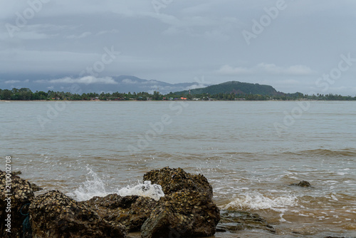 Vászonkép A choppy sea washes against rocky shorelines in front of a distant, verdant coastline under a cloudy sky