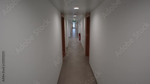 Empty narrow corridor in a modern building lined with wooden doors tiled floor a green emergency exit sign and a red fire extinguisher on the distant wall