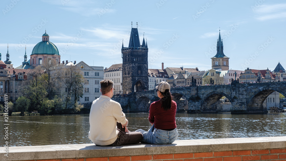 Obraz premium A couple sits peacefully by the Vltava River, admiring beautiful views of Prague. The iconic Charles Bridge and charming buildings create a perfect backdrop for a romantic holiday.
