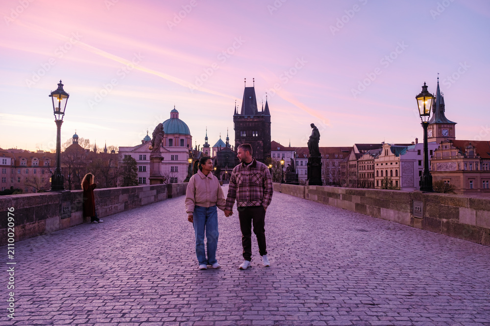Obraz premium A couple walks hand in hand across the iconic Charles Bridge in Prague during a stunning sunset. The vibrant colors reflect the romantic essence of the city, perfect for tourists exploring its charm.