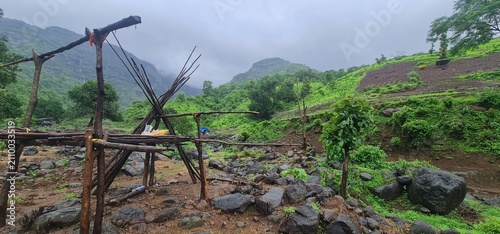 Old Wooden Pole Shelter Frame Situated in Wet Farm Fields Against Lush Green Mountainous Landscape During Monsoon Season