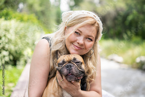 Happy young woman hugging adorable French bulldog while relaxing outdoors in scenic park with warm natural lighting