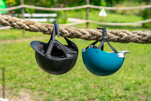Two equestrian riding helmets, one classic black and one blue, hanging from a thick natural rope fence outdoors against a green grass field in daylight.