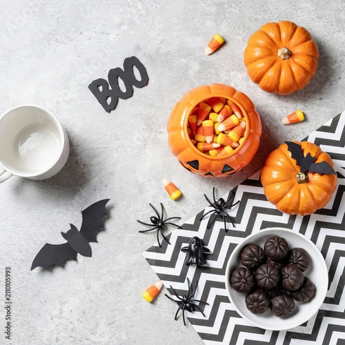 Halloween flat lay featuring a pumpkin candy bowl filled with candy corn, black paper bats, plastic spiders, and a bowl of dark chocolate truffles on a textured gray surface with chevron paper