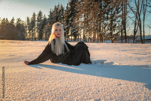 Beautiful blonde girl in black dress sits on white snow in park during winter golden hour sunset.