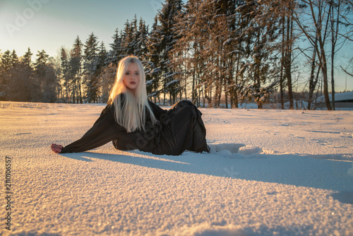 Beautiful blonde girl in black dress sits on white snow in park during winter golden hour sunset.