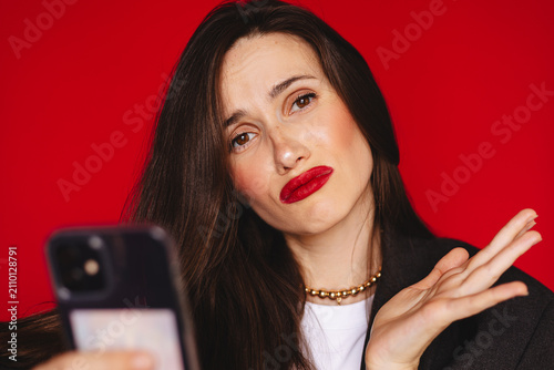 Confused clueless young woman shrugging open palm looking at camera holding smartphone reading strange message wearing dark blazer red lipstick, isolated on studio bright red background