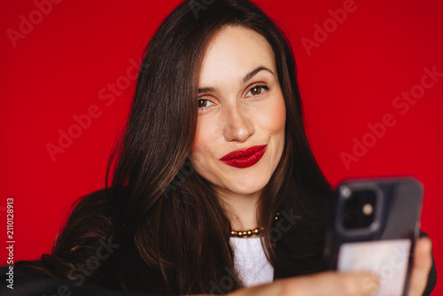 Charming attractive young woman looking at camera with playful smile pouting lips holding smartphone reading message wearing dark blazer red lipstick, isolated on bright studio red background photo