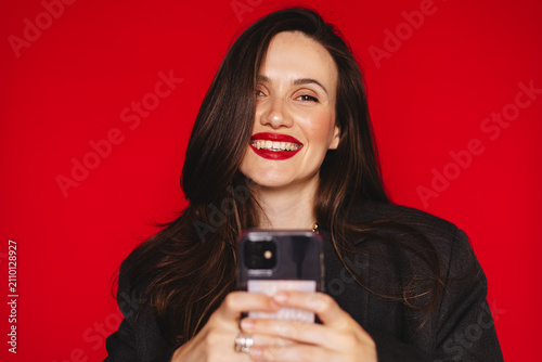 Happy joyful young woman looking at camera with broad smile teeth holding smartphone texting reading message wearing dark blazer red lipstick, isolated on studio bright red background stock photo
