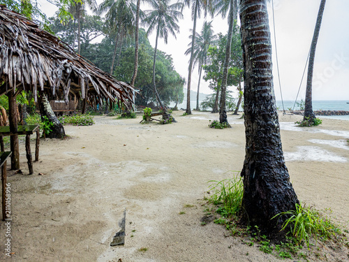 Tropical resort hut featuring a thatched roof on a sandy beach dotted with palm trees during a downpour, offering a quiet, rustic atmosphere in Karimunjawa, Jepara, Jawa Tengah, Indonesia