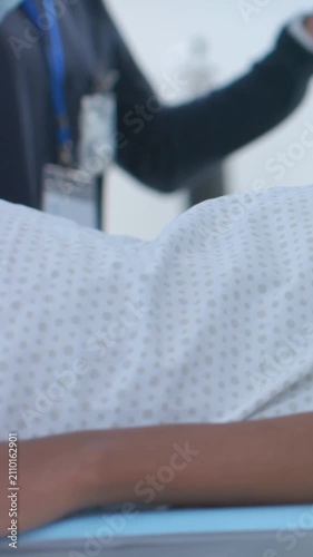 Vertical video: Lying patient wearing dotted gown on bed, worker gesturing to adjust IV pole