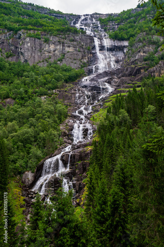 Foto Tall multi-tier waterfall flowing over dark rock with lush green forest on a steep mountainside in summer
