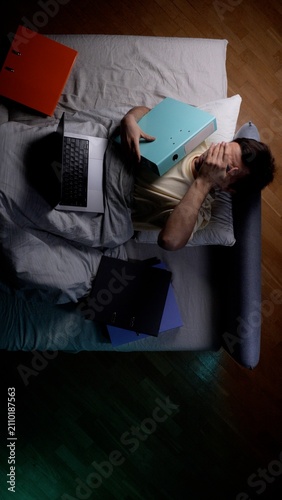 Fotografía Stressed man lying in bed covering his face with one hand, having a folder resti
