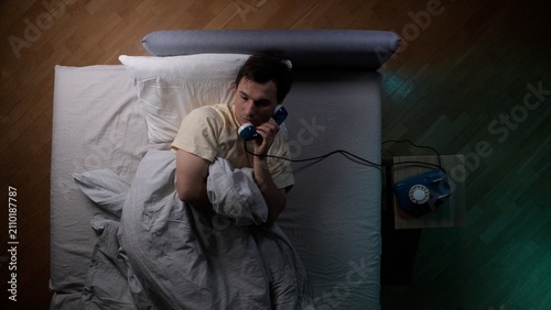 Obraz na płótnie Young man awake in bed at night answering a telephone call, appearing sleepy and