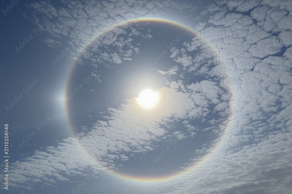 Obraz premium Sun halo with rainbow colors encircles bright sun in blue sky. Altocumulus clouds form pattern around atmospheric phenomenon. Light refraction creates optical illusion during daytime.