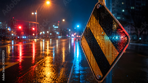 A highly reflective traffic sign illuminated by colorful city lights after the rain; wet asphalt reflecting vibrant red, blue, and yellow hues; shot from a cinematic eye-level perspective on a rainy n
