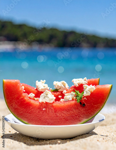 Sliced watermelon and feta cheese on a simple white plate provide a refreshing snack by the sparkling blue sea water.