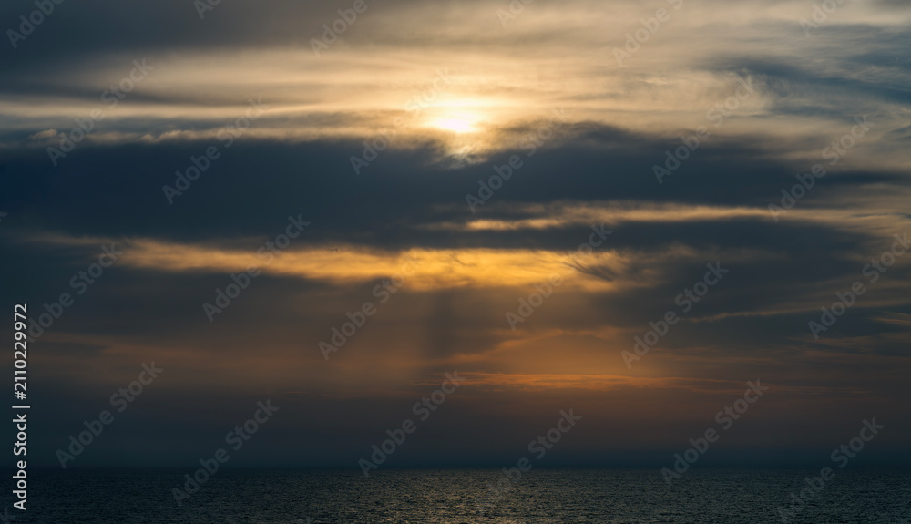 Fototapeta premium Golden sun rays breaking through heavy dark storm clouds over a dark open sea, creating a dramatic, atmospheric, and moody natural light spectacle over calm ocean waters at dusk