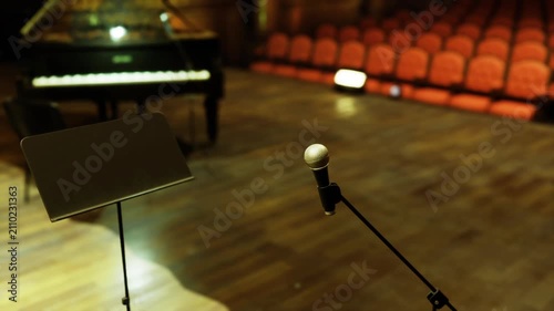 Closeup microphone and music stand on stage, soft spotlight hitting metallic grill, empty auditorium rows and grand piano out of focus suggest pre show calm