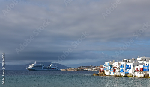 A large cruise ship anchored in the bay off Mykonos, Greece, framed by traditional whitewashed architecture and rustic wooden details. The scene captures the contrast between classic Cycladic design a