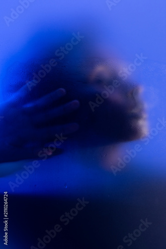 Artistic portrait of young black woman in blue light behind wet frosted glass. 