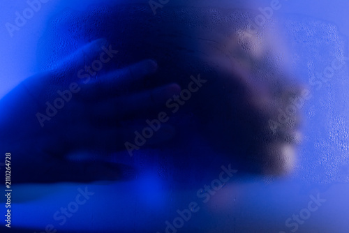 Emotional woman portrait behind wet frosted glass in blue light. Closeup of face and hand 