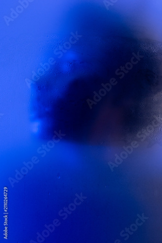 Abstract beauty portrait. Photo of anonymous black woman behind wet matte glass in blue light. 