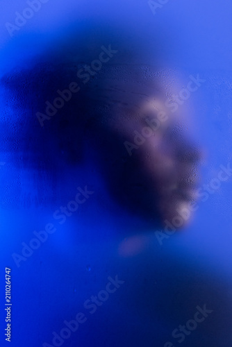 Anonymous beauty. Portrait of young black woman posing in studio with blue light behind wet steamy frost glass.
