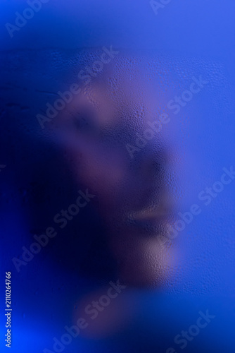 Portrait of young black woman in blue light behind wet matte glass in studio. 