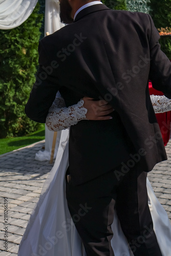 Obraz na plátně Close-up shot of a bride's hand in a lace sleeve tenderly hugging her groom's waist in a black suit