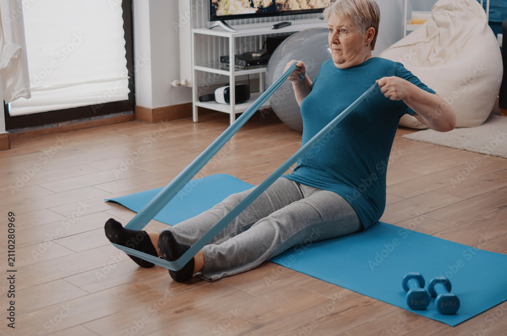 Fototapeta premium Elderly woman exercises with resistance band using an elastic band for stretching, muscle training and gentle rehabilitation during a home fitness routine.