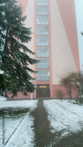 Empty snow covered path to entrance paver walkway leads toward pink highrise lobby with melting patches of snow, evergreen branches overhang sidewalk, soft fog blurs