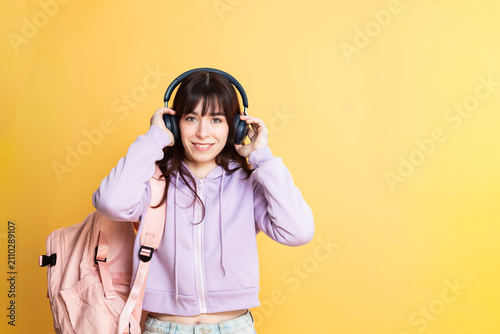 Smiling female student wearing a hooded sweatshirt and a backpack. Putting on wireless headphones. Looking ready for learning and enjoying music against a bright yellow background