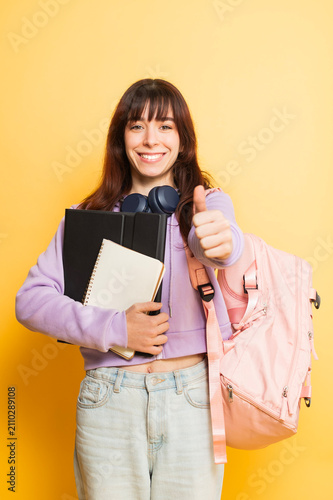 Happy young woman wearing a casual hoodie and jeans, holding academic supplies and a pink backpack, smiling and showing a thumbs up gesture, ready for school or university
