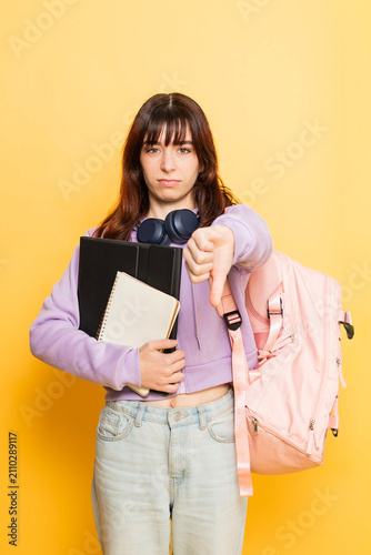 Young female student holding school books and a backpack, expressing her dislike for returning to education by giving a thumbs down gesture against a yellow background