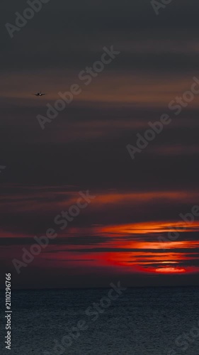 Airplane flying across dark moody sky over calm ocean at dramatic red sunset. Vertical orientation, wide shot, evening twilight atmosphere with colorful clouds.