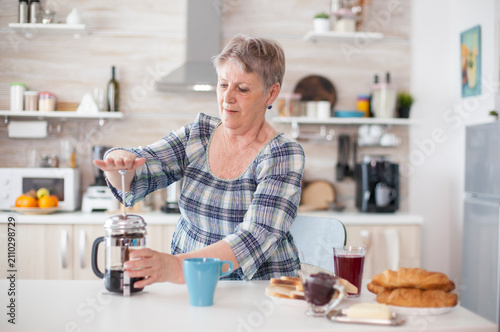 Fotografi Senior woman makes coffee with a French press in the kitchen during breakfast, preparing a fresh caffeinated drink for the morning, inside a calm morning kitchen routine