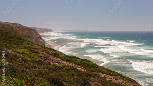 Majestic view of the vicentina coast in portugal, showing powerful ocean waves rolling onto a sandy beach at the bottom of a lush green cliff on a sunny day in the algarve region