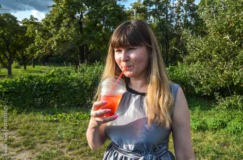 Young woman sips a bright red iced beverage through a straw while standing in a green park, enjoying a warm summer afternoon and a relaxed, casual outdoor moment