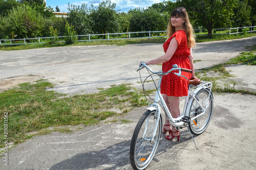 Woman in a red polka dot dress stands beside a white bicycle on a sunlit country road, conveying relaxed summer style, outdoor leisure and a nostalgic, carefree mood