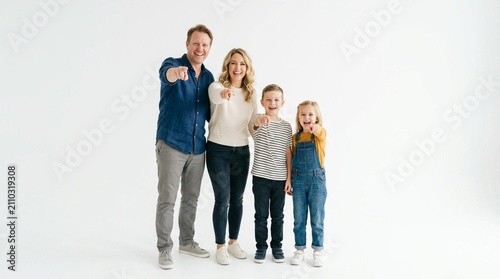 Happy family of four, parents and children, standing together in bright white studio, all pointing forward with enthusiasm, wearing casual autumn clothes, perfect for family and lifestyle content.