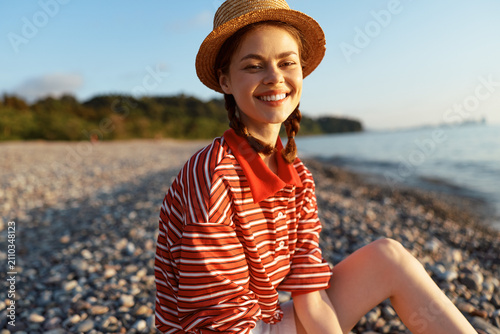 Fotografie Young woman in a red striped sweater and straw hat sits on a pebbly beach, smiling at the camera as golden hour light warms the scene, creating a relaxed vibe