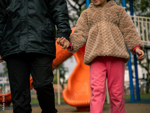 Happy child holds adult's hand at playground.nAdult wears black raincoat