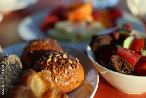 A close-up of assorted breakfast foods including seeded rolls, pastries, and a fresh tomato salad with fruit in warm morning sunlight.