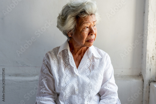 Fotografi Elderly woman with white hair gazing out a window, wearing a white blouse