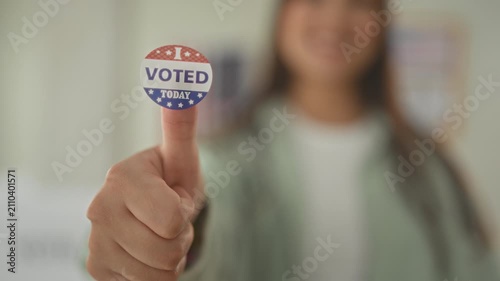 Young hispanic woman holds 'i voted today' sticker on raised thumb in a polling building, smiling and offering the sticker toward camera; civic pride.
