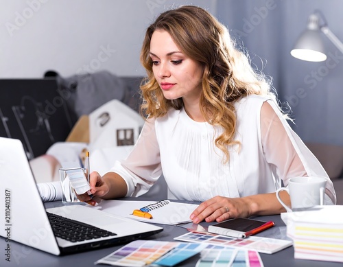 A woman with wavy hair works at a desk, looking at a notepad and laptop. Home decor models are in the background