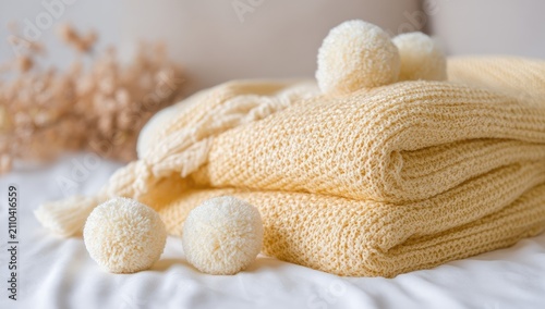 Close-up of folded yellow knitted blanket with pom-poms on a white bed. Soft, cozy, and inviting feel