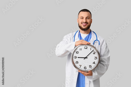 Smiling male doctor in a white coat and stethoscope holding a large wall cloc...